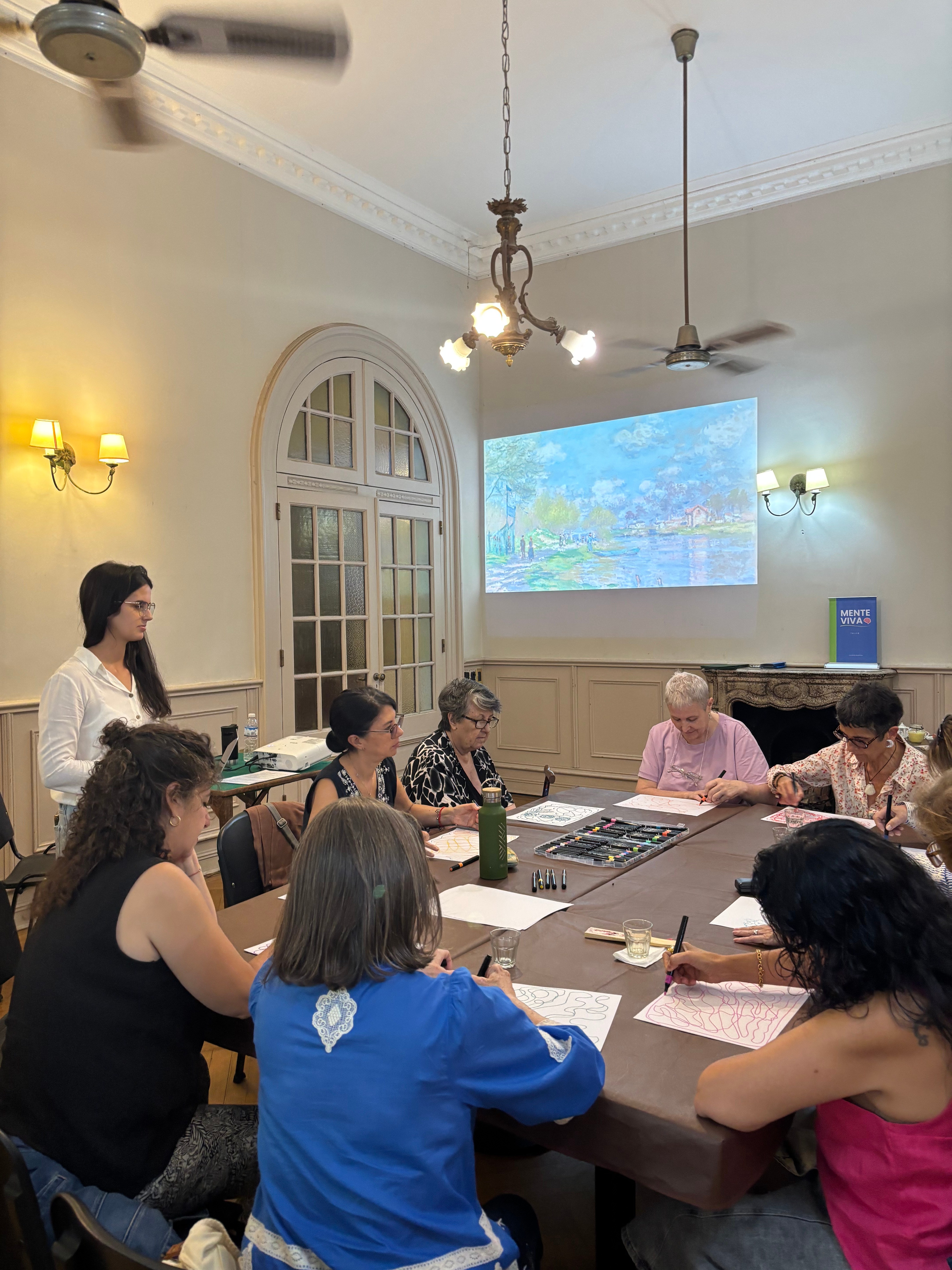 Participantes trabajando alrededor de la mesa durante el taller de arteterapia, con la tallerista de pie y una obra impresionista proyectada en la pared junto al banner de Mente Viva.