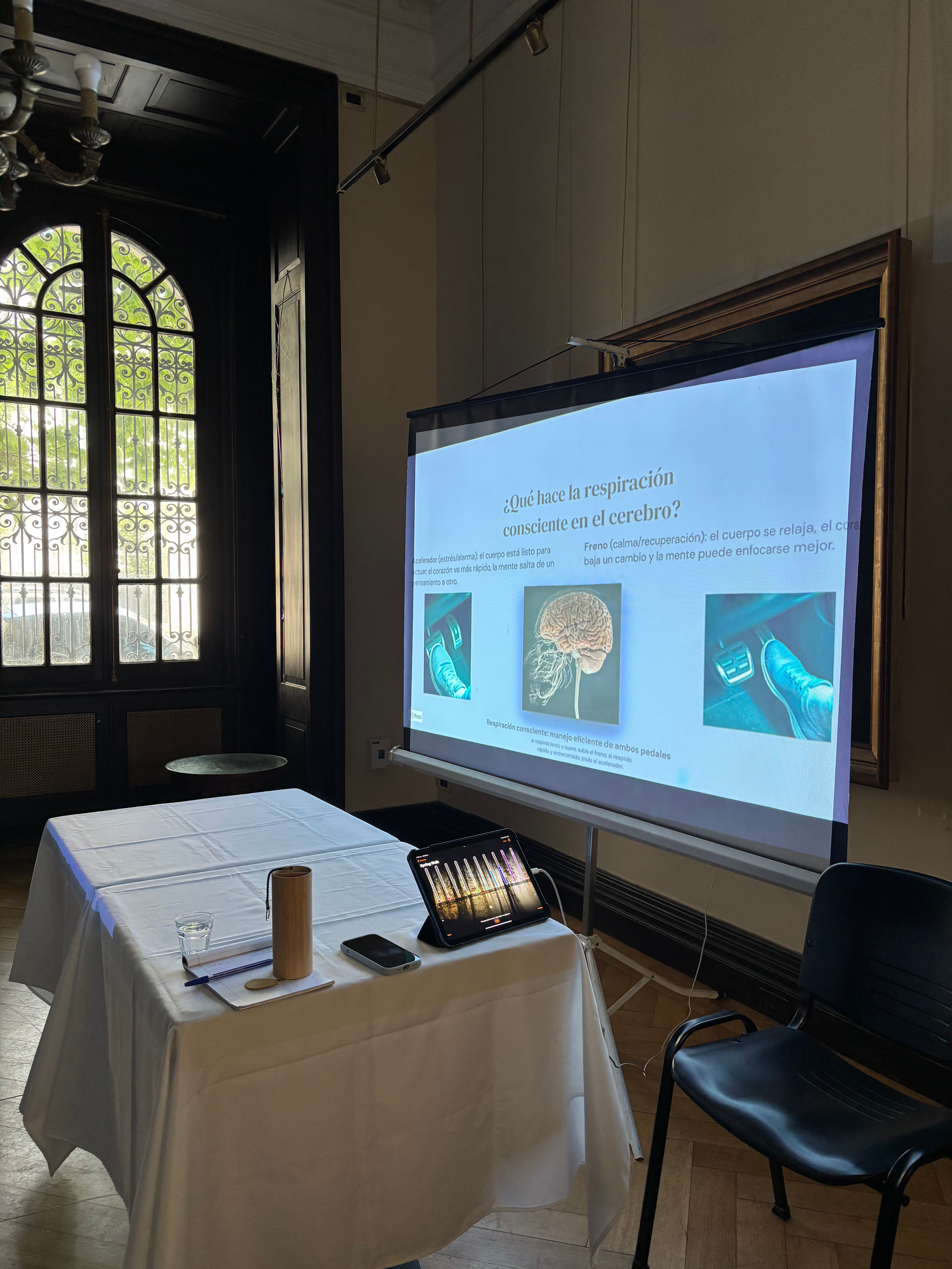 Sala del encuentro de respiración consciente con una diapositiva proyectada '¿Qué hace la respiración consciente en el cerebro?' y una mesa con materiales del taller.